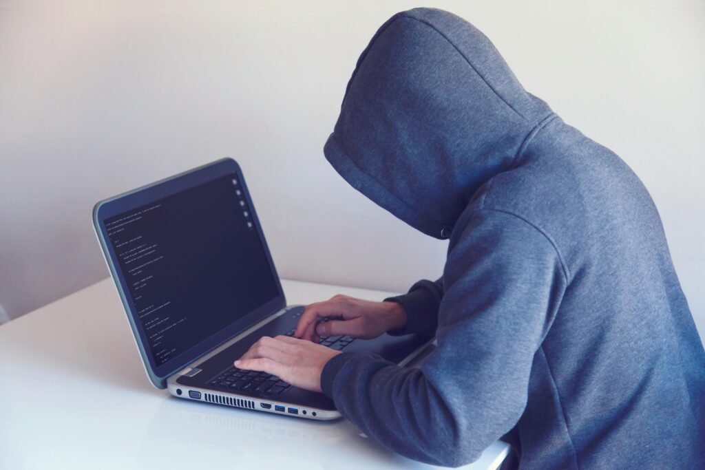 Side view of unrecognizable hacker in hoodie sitting at white table and working remotely on netbook in light room near wall