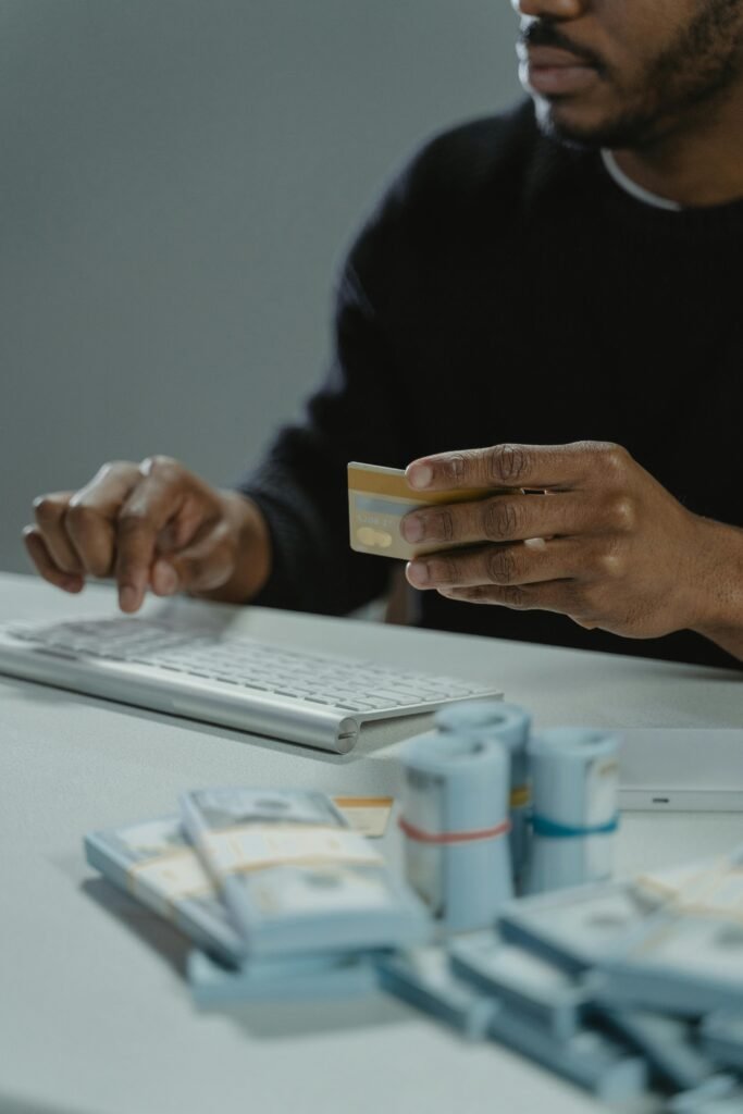 Person typing on keyboard holding credit card with stacks of cash nearby, indicative of online transactions.