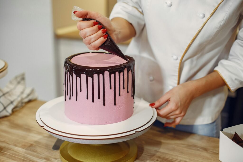 Anonymous female pastry chef in white uniform standing near counter and preparing yummy cake with chocolate drips from pastry bag in kitchen