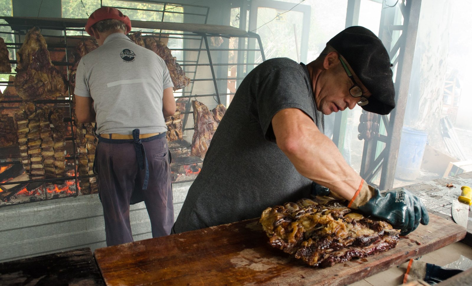 Two men preparing meat for barbecue on an outdoor grill with smoke rising.