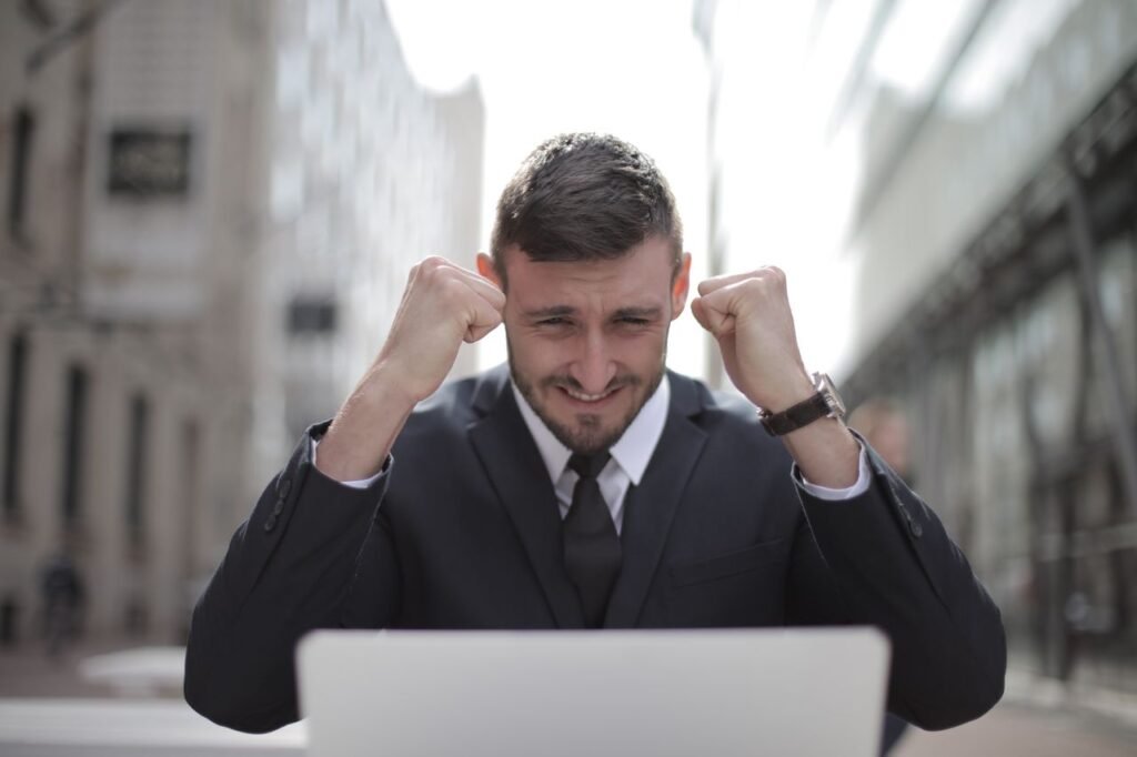 Como Encontrar Vagas de Emprego: Os Melhores Sites para Procurar Trabalho em 2026 A joyful businessman in a suit triumphantly celebrates success on his laptop outdoors.
