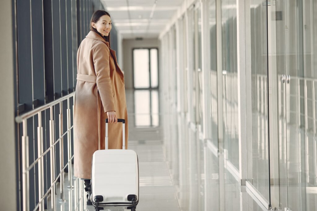 Back view full length of young cheerful female passenger in trendy beige coat walking along airport airbridge and looking at camera over shoulder